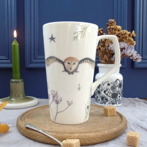 May include: A white ceramic mug with a tall, slender design features a hand-painted illustration of an owl in flight against a backdrop of delicate floral sprigs and a single star. The mug has a large handle and is perfect for enjoying a warm beverage.