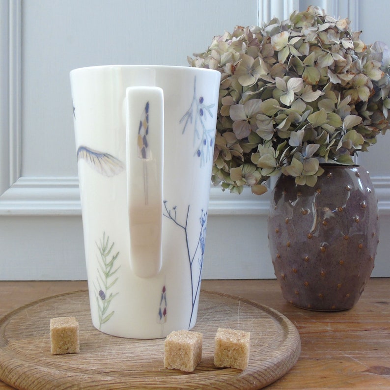 May include: A tall white ceramic mug with a floral design and a handle. The mug is sitting on a wooden surface with three brown sugar cubes.