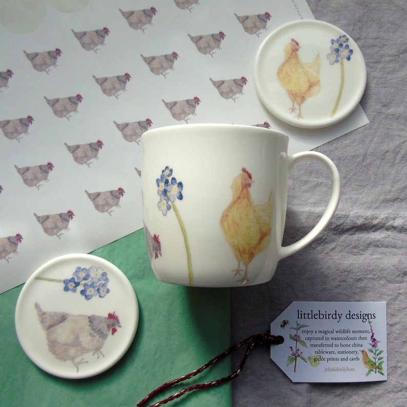 May include: A white ceramic mug with a yellow chicken and blue forget-me-not flowers painted on it. The mug is next to a coaster with a gray chicken and blue forget-me-not flowers. The image also includes a sheet of stickers with gray chickens and a tag with the text "littlebirdy designs".