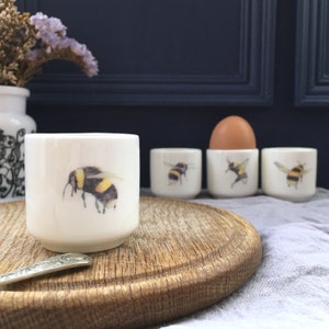 May include: Four white ceramic egg cups with a bumble bee design. One egg cup has a boiled egg inside. The egg cups are on a wooden surface.