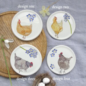 May include: Four white ceramic coasters with different designs featuring chickens and forget-me-not flowers. The coasters are arranged on a wooden surface. The text "design one", "design two", "design three", and "design four" is written below each coaster.