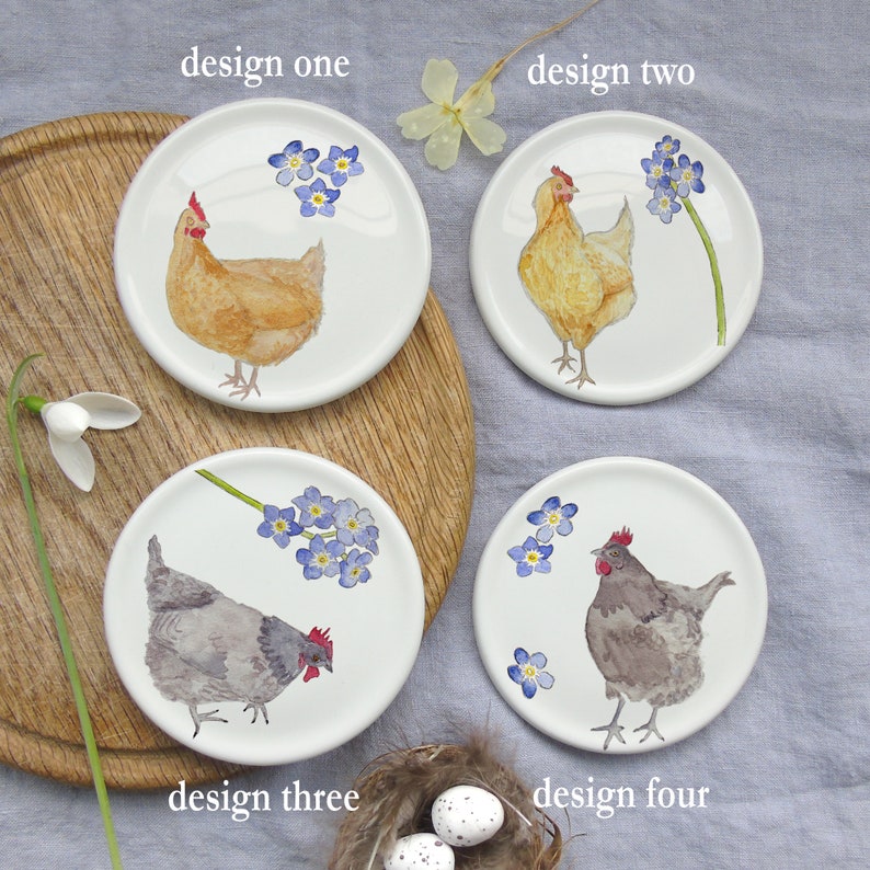 May include: Four white ceramic coasters with different designs featuring chickens and forget-me-not flowers. The coasters are arranged on a wooden surface. The text "design one", "design two", "design three", and "design four" is written below each coaster.