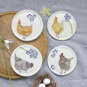 May include: Four white ceramic coasters featuring watercolour illustrations of chickens and blue forget-me-nots. The chickens are in shades of brown, yellow, and grey. The coasters are arranged on a wooden tray and grey fabric.
