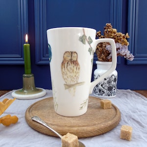 May include: A white ceramic mug with a square handle, decorated with a watercolour illustration of a sleeping owl and ivy. A green candle in a ceramic holder is next to the mug. The mug is on a wooden tray with sugar cubes.