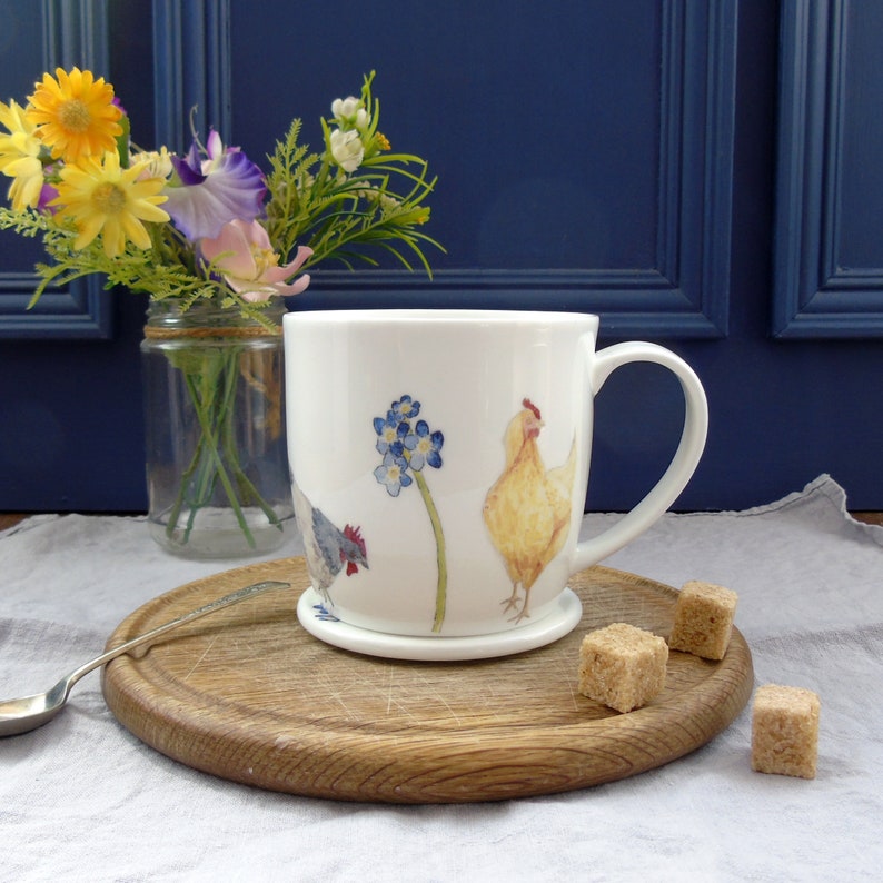 May include: White ceramic mug with a hand-painted design of chickens and forget-me-not flowers. The mug is on a wooden coaster and there are sugar cubes on a wooden surface.