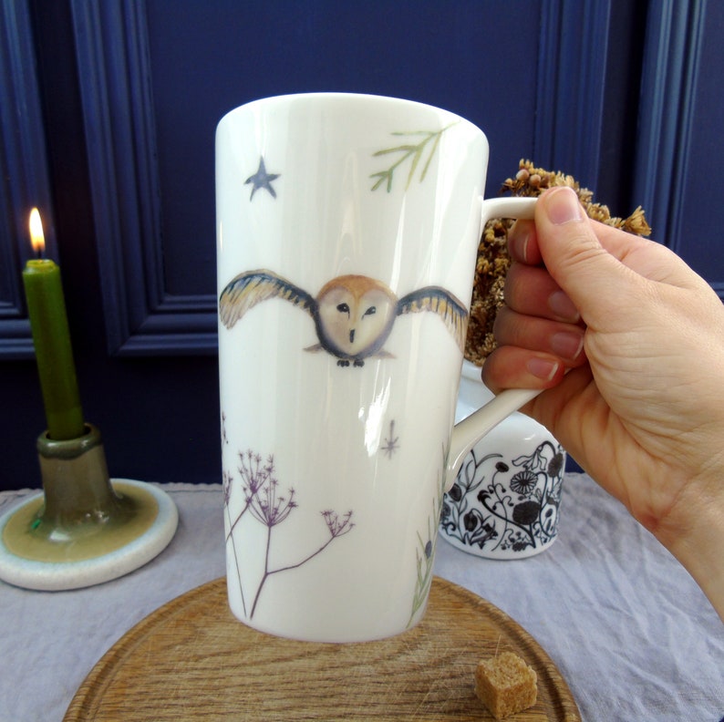 May include: White ceramic mug with a tall, cylindrical shape. The mug features a hand-painted design of an owl in flight, surrounded by delicate floral sprigs and stars.
