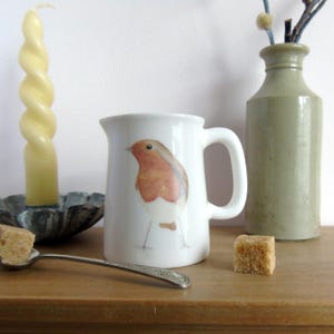 May include: White ceramic pitcher with a handle, featuring a watercolor illustration of a robin. A twisted yellow candle sits in a silver dish. A silver spoon holds a sugar cube, and a green vase is in the background.