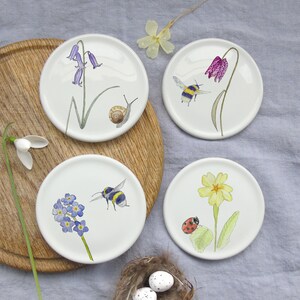 May include: Set of four white ceramic coasters with floral and insect designs. Each coaster features a different design: bluebells and a snail, a bee and a pink flower, forget-me-nots and a bee, and a yellow primrose with a ladybug.