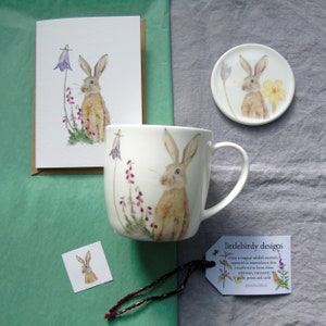 May include: A set of watercolor illustrations featuring a brown hare in a field of wildflowers. The set includes a mug, a coaster, a card, and a small sticker. The illustrations are printed on bone china and are available in a variety of sizes.