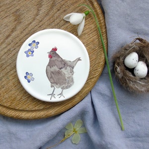 May include: A white ceramic plate with a hand-painted design of a brown hen with a red comb and blue forget-me-not flowers. The plate is on a wooden surface.