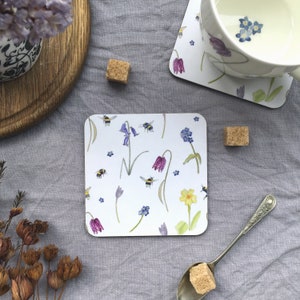May include: A set of four square coasters featuring a floral design. The coasters have a white background with a repeating pattern of bluebells, bees, and other wildflowers. The coasters are made from a durable material and are ideal for protecting your table from heat and spills.