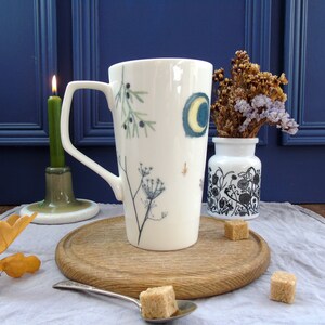 May include: A white ceramic mug with a tall, slender design. The mug features a hand-painted design of a crescent moon, stars, and botanical elements in shades of blue, green, and yellow. The mug is sitting on a wooden surface.