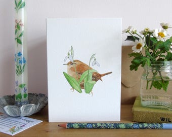 May include: A watercolor illustration of a brown wren perched on a snowdrop plant with white flowers. The bird is facing the right side of the image. The illustration is printed on a white card.