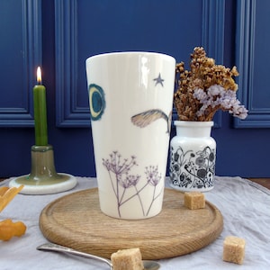 May include: A white ceramic mug with a whimsical design featuring a bird in flight, a crescent moon, a star, and delicate floral sprigs. The mug is on a wooden surface with sugar cubes nearby.