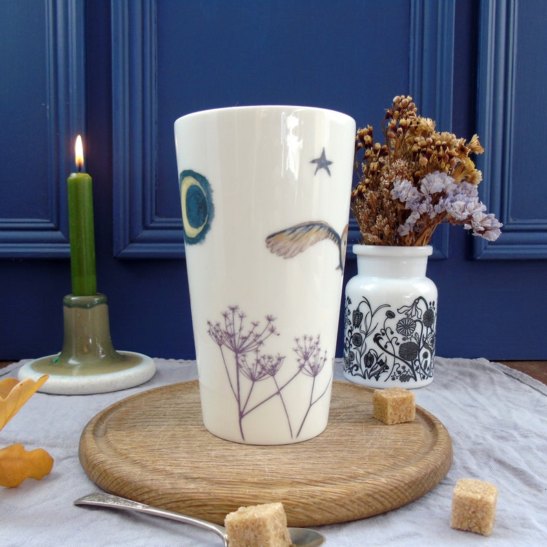May include: A white ceramic mug with a whimsical design featuring a bird in flight, a crescent moon, a star, and delicate floral sprigs. The mug is on a wooden surface with sugar cubes nearby.