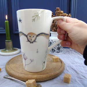May include: White ceramic mug with a brown and white barn owl flying in front of a white background with green leaves and a few stars. The mug has a white handle.