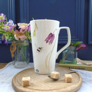 May include: White ceramic mug with a floral design featuring a bee, a pink flower, and yellow flowers. The mug has a handle and is sitting on a wooden surface with sugar cubes and a spoon.