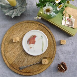 May include: A white ceramic saucer with a hand-painted illustration of a robin and a snowdrop flower. The saucer is on a wooden board with sugar cubes and a spoon.