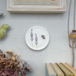 May include: A white ceramic coaster with a painted design of lavender and a bumblebee. The coaster is round and small, suitable for placing under a mug or glass. The design is a watercolor style.