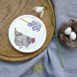 May include: A white ceramic plate with a gray hen and a cluster of blue forget-me-not flowers painted on it.
