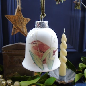 May include: A white ceramic bell ornament with a silver top and a painted image of a brown wren perched on a green leaf. The bell is hanging from a green ribbon.