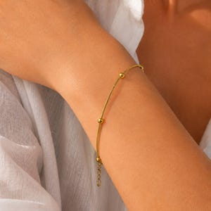 May include: A gold-toned bracelet featuring a thin, flexible snake chain design accented with small, evenly spaced spherical beads. The piece includes a short adjustable extender chain with a small circular link at the end, providing a secure fit.