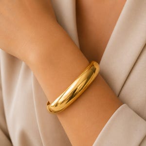 May include: A gold-coloured bangle bracelet. The bracelet is a solid, rounded band, reflecting light. The jewellery is worn on a person's arm, with a neutral-toned background.