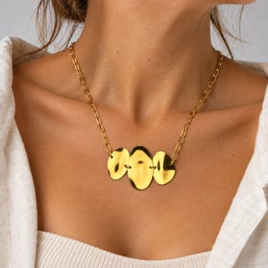 May include: A gold-toned necklace featuring a chain with elongated oval links and a central pendant composed of three smooth, reflective, irregular oval-shaped metal discs connected horizontally in a row against a light-coloured fabric.