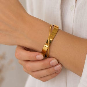 May include: A gold-coloured bangle bracelet with a cross-over design. The bracelet is made of a shiny, reflective metal. The image shows the bracelet on a person's wrist, with a white shirt in the background.
