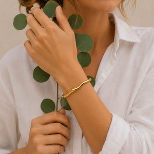 May include: A gold-coloured bracelet with a wavy design. The bracelet is worn on the wrist. The image also shows a person holding a green leafy plant and wearing a white button-down shirt.