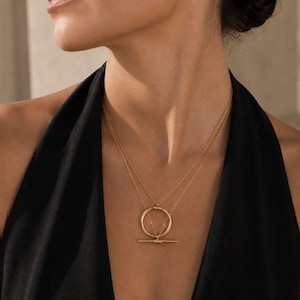 Può includere: Set di collane dorate con due delicate catene. La catena superiore ha un ciondolo circolare, e la catena inferiore ha un ciondolo a barra orizzontale. I gioielli sono esposti su un top nero.