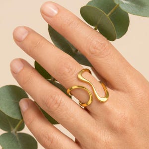 May include: A gold ring with a distinctive, flowing design. The ring has a curved, open shape that wraps around the finger. The jewellery is displayed on a hand with a neutral background and greenery.