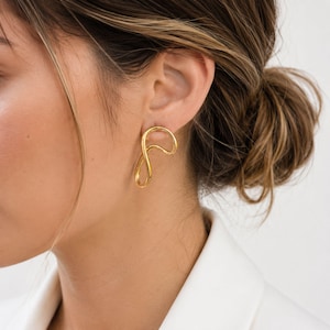May include: A pair of gold-coloured, abstract-shaped earrings. The earrings have a flowing, looped design, and are worn on the ear. The earrings are a bright, shiny gold colour.