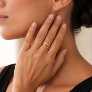 May include: A set of three gold-toned rings worn on a finger. The top ring is a smooth, thin band, the middle ring features a textured, hammered surface, and the bottom ring is a plain, thin band. The rings are stacked together on the finger.