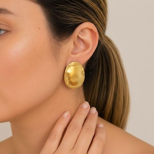 May include: A close-up of a gold-coloured, oval-shaped earring. The earring has a textured surface and is worn on the ear. The image shows a portion of a person's face, ear, and neck.