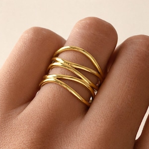 May include: A gold ring with a multi-layered design. The ring features several curved bands that cross over each other, creating a woven effect. The ring is worn on a finger, showcasing its intricate and modern style.
