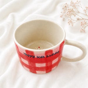 May include: A ceramic mug featuring a red and white chequered pattern on the exterior and the text "Happy 30th birthday" written in black. The interior is cream-coloured and contains a small, sculpted figure at the bottom of the basin.