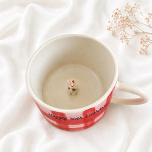 May include: A ceramic mug with a red and white checkered pattern on the exterior and the text Happy 30th birthday written in black. Inside the mug, a small, sculpted white dog figure wearing a red and white polka dot party hat sits at the base.