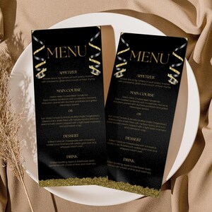May include: Two black menu cards with gold text and accents, featuring a menu for appetizers, main courses, desserts, and drinks. The cards are on a white plate, with a beige background and a sprig of dried grass.