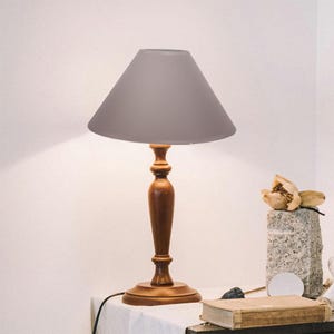 May include: A table lamp featuring a smooth, medium-brown wooden base with a turned, spindle-style column and a wide, circular pedestal. The lamp is topped with a solid, light-grey conical shade that directs light downwards from the centre.