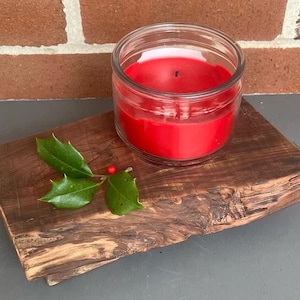 Puede incluir: Una vela roja en un tarro de cristal transparente se encuentra sobre una tabla de madera rústica, con hojas verdes de acebo y bayas rojas. La mecha de la vela es visible. La tabla de madera tiene un borde natural sin terminar.