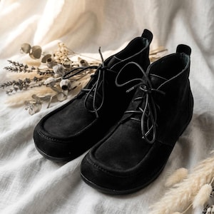 May include: A pair of black suede ankle boots with a lace-up design. The boots have a rounded toe and a low heel. The boots are displayed on a white fabric surface with dried flowers in the background.