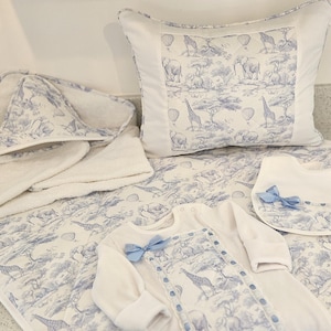 May include: A set of baby items featuring a blue and white toile print with elephants, giraffes, and hot air balloons. The collection includes a quilted pillow, a hooded towel, a long-sleeved onesie with a blue bow, and a matching bib with a bow.
