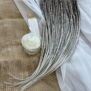 Könnte beinhalten: Ein Bündel grauer und weißer Kunst-Dreadlocks. Die Dreadlocks sind auf einer strukturierten, beigen Oberfläche mit einer weißen Feder und einem kleinen, durchsichtigen Glasbehälter mit Deckel angeordnet. Die Dreadlocks haben einen Farbverlauf.