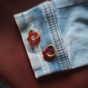 Peut inclure: Une paire de boucles d'oreilles créoles dorées avec un motif floral rouge pailleté. Les boucles d'oreilles sont présentées sur un tissu en jean, avec un fond en cuir bordeaux texturé.