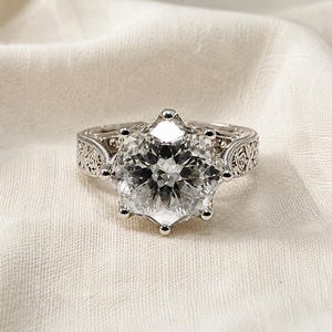May include: A silver ring with a large, clear, faceted gemstone. The ring has an ornate, filigree design on the band and around the gemstone setting. The ring is displayed on a white, textured surface.