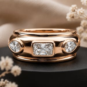 Peut inclure: Une bague en or rose avec une pierre centrale rectangulaire taille radiant, encadrée par deux pierres plus petites en forme de poire. Le métal présente une finition polie et réfléchissante avec un profil large et des sertis clos.