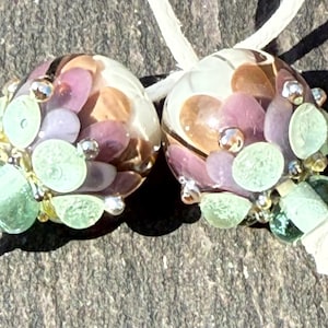 May include: Two spherical glass beads featuring a clear base with embedded purple, brown, and light green circular patterns. Each bead is accented with small silver-toned metallic spheres and is threaded onto a thin white cord for display.