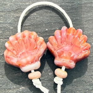 Puede incluir: Dos cuentas de vidrio con forma de conchas de vieira en tonos rosa, naranja y blanco, cada una con una textura acanalada. Las cuentas están ensartadas en un cordón blanco, con una cuenta espaciadora pequeña, lisa y de color rosa claro.