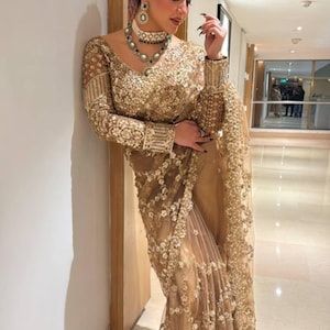 Puede incluir: Un sari dorado con intrincados bordados y adornos. El sari presenta mangas largas y un escote en V, con un collar gargantilla y pendientes a juego. La tela parece ser transparente, con detalles florales.
